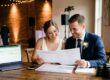 A happy engaged couple, sitting at a rustic table, points to a calendar marked 'DJ BOOKED!' while reviewing wedding planning timeline documents.